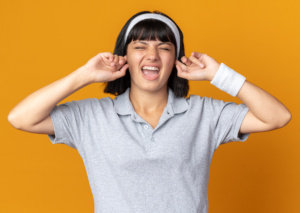 young fitness girl wearing headband closing ears with fingers with annoyed expression standing over orange background